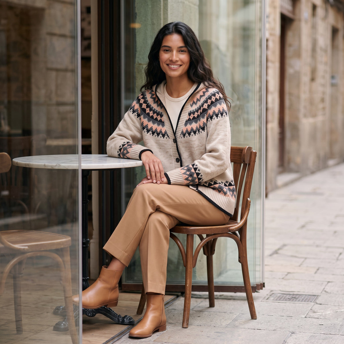 Mujer sentada en la terraza de un café con un cárdigan jacquard nórdico con capucha beige
