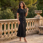 Essyro black ribbed knit maxi dress with gold buttons, short sleeves, and a ruffled hem worn by a woman outdoors.