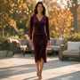Essyro burgundy velvet midi dress with long sleeves, V-neck, and draped waist, worn by a woman on a sunny patio.