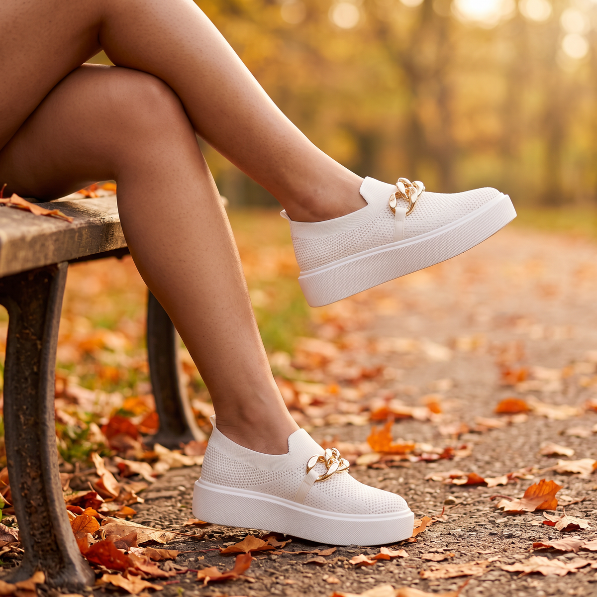Mujer sentada en un banco de parque en otoño con zapatillas slip-on blancas con cadena dorada